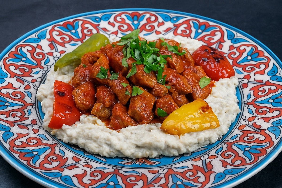 Ароматно ястие Али Назик кебап – нежни късчета агнешко в доматен сос върху крем от патладжан и кисело мляко, поднесени в чиния с турски орнаменти и печени чушки.