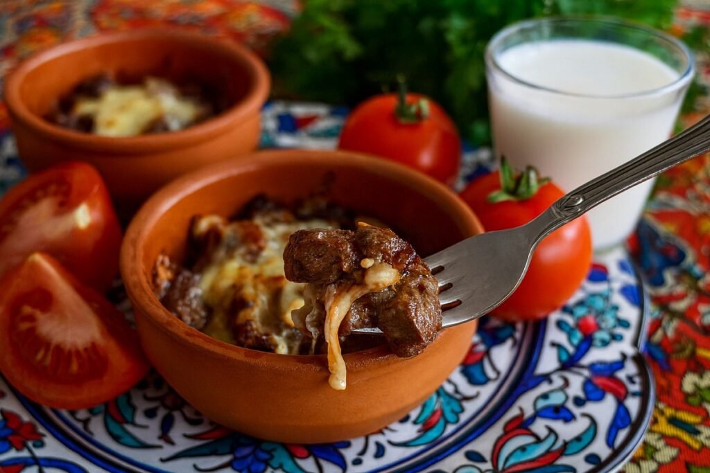 Хапка сочна ориенталска гювеч-кавръма, вдигната на вилица над глинено гювече, с разтопено сирене, пресни домати и чаша айрян върху шарена турска покривка.