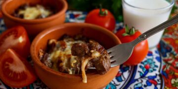 Хапка сочна ориенталска гювеч-кавръма, вдигната на вилица над глинено гювече, с разтопено сирене, пресни домати и чаша айрян върху шарена турска покривка.