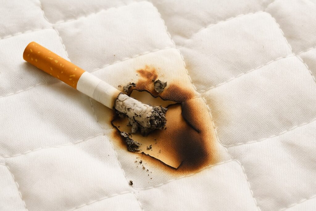 Изгоряла цигара върху бяло одеяло, оставила обгорено петно и следи от пепел – илюстрация за пожар, причинен от небрежност при пушене в леглото.