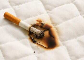 Изгоряла цигара върху бяло одеяло, оставила обгорено петно и следи от пепел – илюстрация за пожар, причинен от небрежност при пушене в леглото.