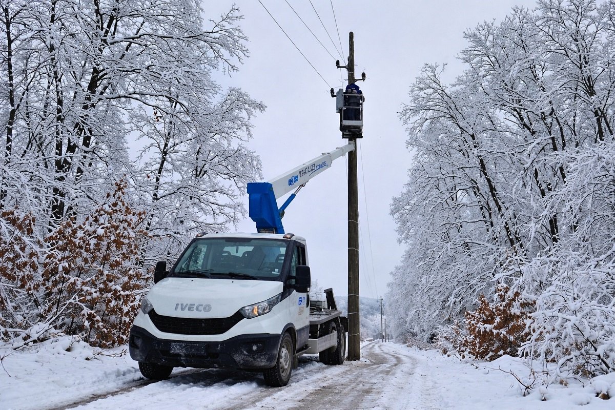 Електротехник отстранява авария по електропровод върху стълб с автовишка и сервизен камион на заснежен път през зимата