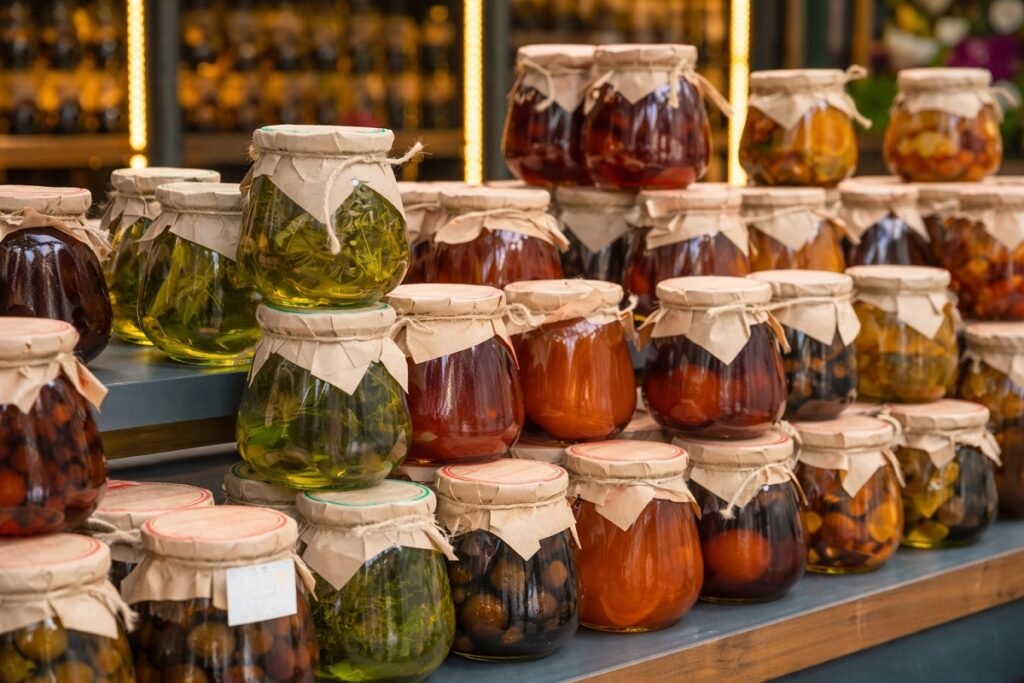 Буркани с домашно приготвени сладка, конфитюри и туршии, подредени на щанд на фермерски пазар, с различни цветове и натурална визия.