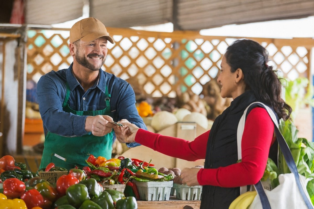 Продавач на фермерски пазар подава рестото на клиентка, двамата се усмихват над щанд с пресни зеленчуци в уютна пазарна среда.