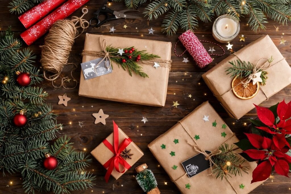 Коледни подаръци, опаковани в кафява крафт хартия, декорирани с канап, зелени клонки, сушени портокалови резени и червени акценти, подредени върху дървена повърхност с празнична украса и свещ.