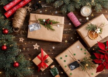 Коледни подаръци, опаковани в кафява крафт хартия, декорирани с канап, зелени клонки, сушени портокалови резени и червени акценти, подредени върху дървена повърхност с празнична украса и свещ.