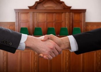 Ръкостискане между две страни в съдебна зала – символ на постигнато споразумение чрез съдебна медиация, на фона на съдийската банка и зелени столове.