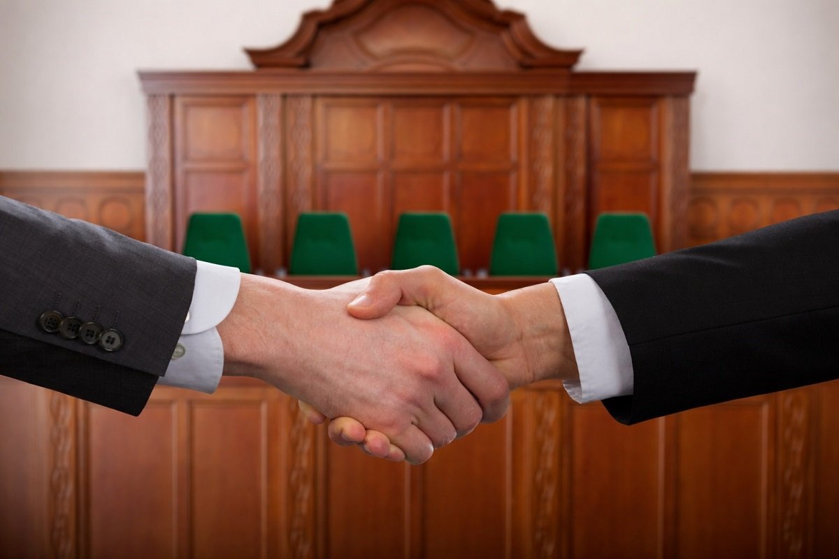 Ръкостискане между две страни в съдебна зала – символ на постигнато споразумение чрез съдебна медиация, на фона на съдийската банка и зелени столове.