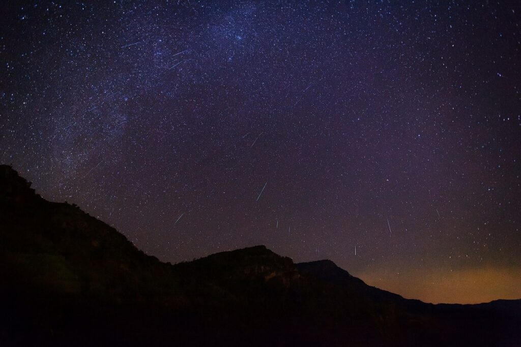„Нощно небе над планински силуети с ярко падащи метеори от потока Геминиди, видими като светли следи върху звездния фон
