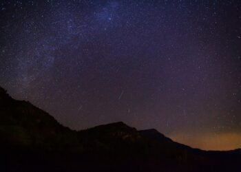 „Нощно небе над планински силуети с ярко падащи метеори от потока Геминиди, видими като светли следи върху звездния фон