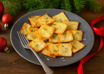 Хоризонтално коледно изображение на подредени хрупкави златисти картофени квадратчета в чиния, поръсени с билки и морска сол, поставени върху дървена повърхност с елхови клонки и червени коледни украшения на заден план.