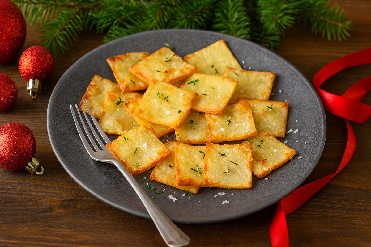 Хоризонтално коледно изображение на подредени хрупкави златисти картофени квадратчета в чиния, поръсени с билки и морска сол, поставени върху дървена повърхност с елхови клонки и червени коледни украшения на заден план.