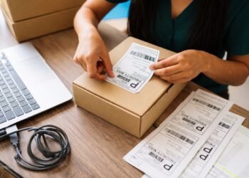 Опаковане на малък колет с наложен платеж – ръце поставят товарителница върху картонена кутия на работно бюро с лаптоп и документи за доставка