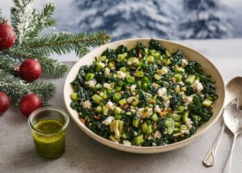 Хоризонтално зимно изображение на голяма купа със зелена салата с кейл, праз, ядки и зелено дресинг, поставена върху светла повърхност. До нея има метални прибори, бурканче с дресинг и коледни елементи като заснежени елхови клонки и червени топки, със зимен пейзаж в размит фон.