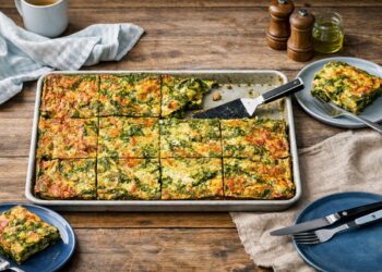 Домашно приготвена спаначена фритата със сирене, нарязана на квадрати в метална тава, сервирана върху дървена маса с чинии, прибори и кухненски текстил, осветена от естествена дневна светлина.