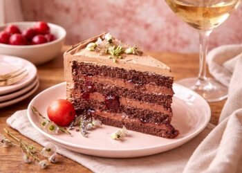 Парче домашна шоколадова торта с крем и вишни, поднесено върху чиния на дървена маса с елементи на домашен уют и мека естествена светлина.