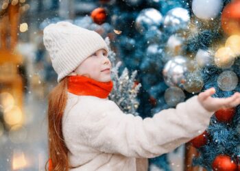 Дете с бяла шапка и шал пред украсена коледна елха в снежна зимна атмосфера.