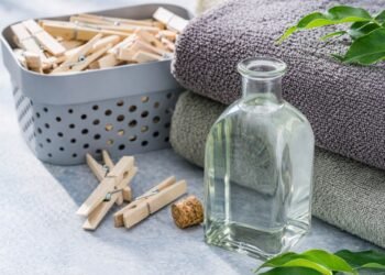 Стъклена бутилка с оцет без етикет до навити хавлиени кърпи, кошница с дървени щипки и зелени листа върху светла повърхност, хоризонтален кадър с натурална светлина.