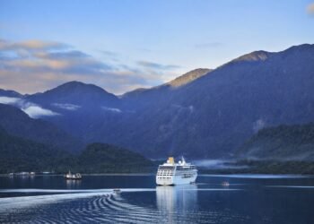 Круизен кораб в спокоен фиорд, заобиколен от зелени планини и отразен във водата при меко утринно осветление