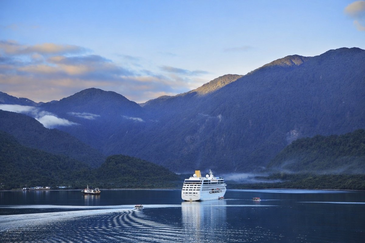 Круизен кораб в спокоен фиорд, заобиколен от зелени планини и отразен във водата при меко утринно осветление