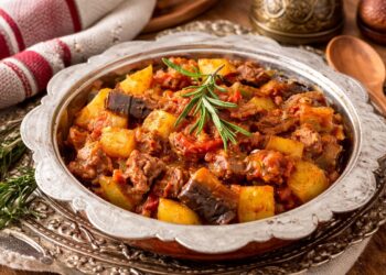 Апетитен турски гювеч с крехко месо, картофи, патладжан и доматен сос, поднесен в декоративен съд, гарниран с пресен розмарин, заснет хоризонтално при топла светлина.