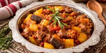 Апетитен турски гювеч с крехко месо, картофи, патладжан и доматен сос, поднесен в декоративен съд, гарниран с пресен розмарин, заснет хоризонтално при топла светлина.