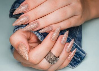 лги стилетни нокти в розово-златист глитер, елегантен маникюр с блясък, ръце върху деним с декоративен сребърен пръстен, модерен и луксозен стил.