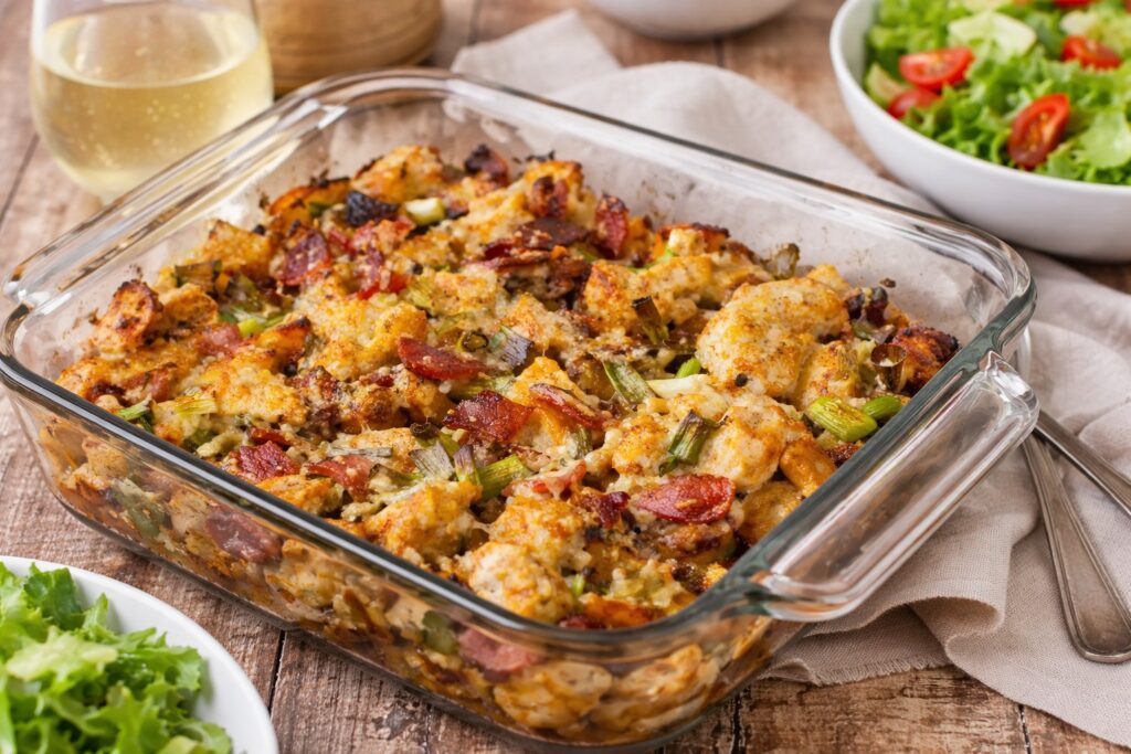 Фурнено пилешко с картофи, разтопен чедър и хрупкав бекон в стъклена тава, поднесено на дървена маса с чаша бяло вино и свежа зелена салата, хоризонтален кадър без надписи.