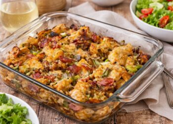 Фурнено пилешко с картофи, разтопен чедър и хрупкав бекон в стъклена тава, поднесено на дървена маса с чаша бяло вино и свежа зелена салата, хоризонтален кадър без надписи.