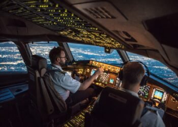 Пилотска кабина на пътнически самолет по време на нощен полет, с двама пилоти и осветени навигационни уреди над градски светлини