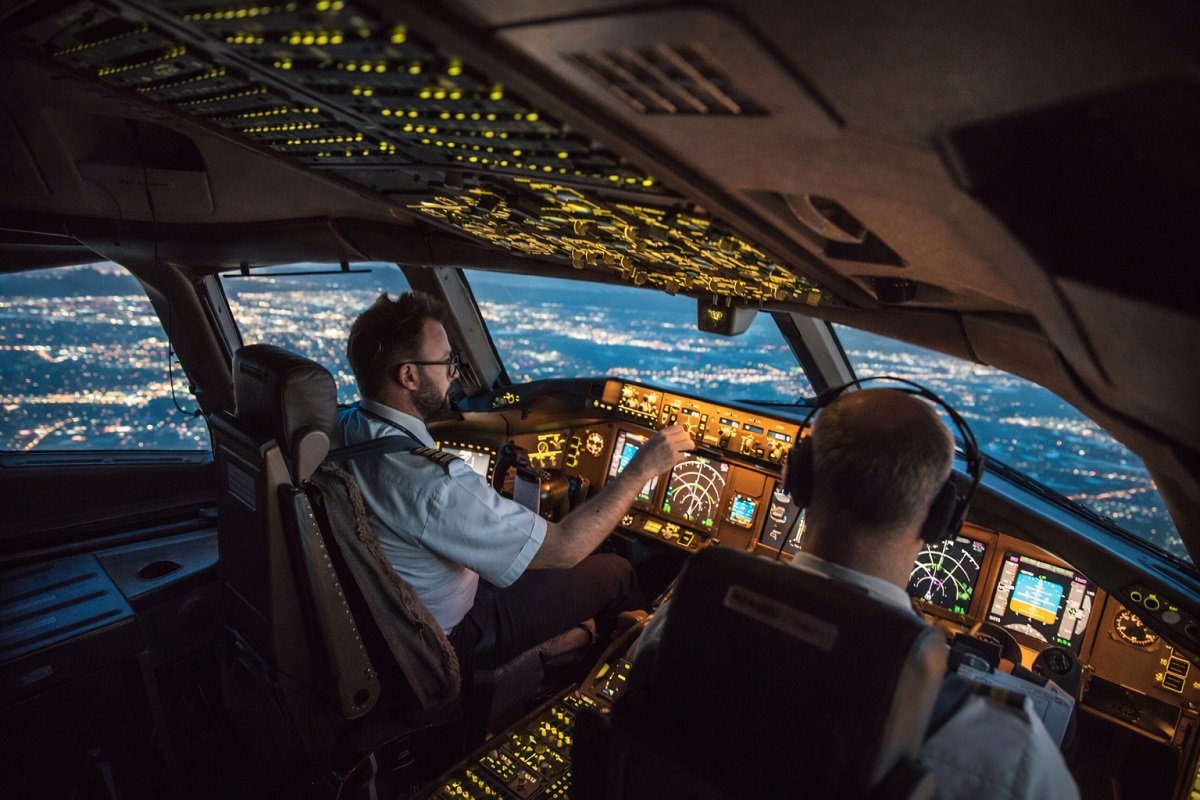 Пилотска кабина на пътнически самолет по време на нощен полет, с двама пилоти и осветени навигационни уреди над градски светлини