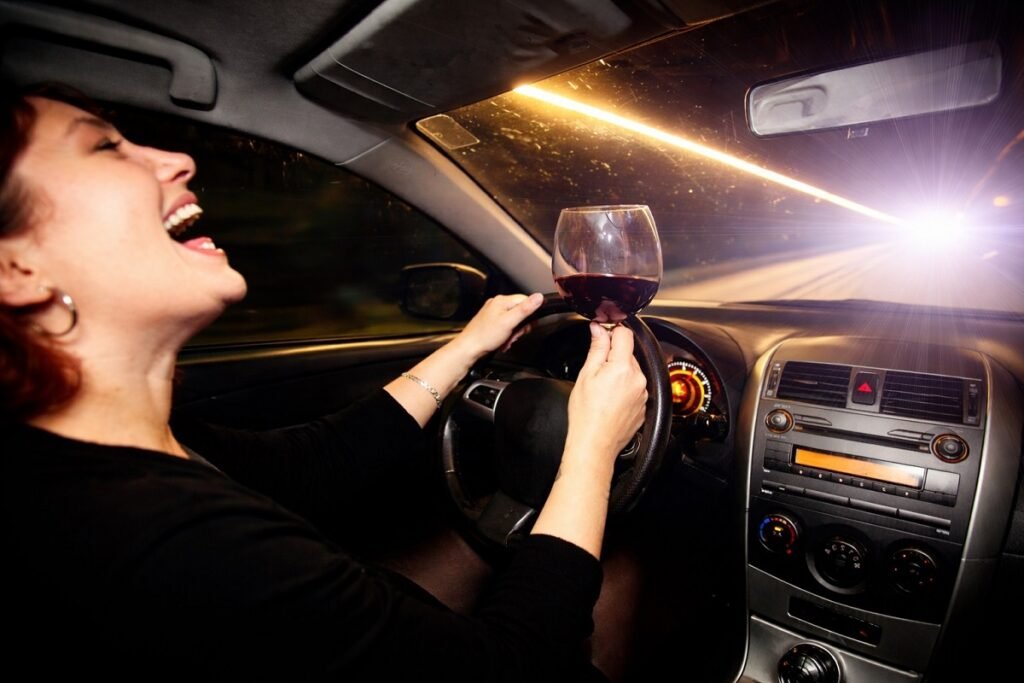 Жена зад волана на автомобил през нощта, държи чаша с вино, силни светлини от насрещното движение и динамична пътна обстановка