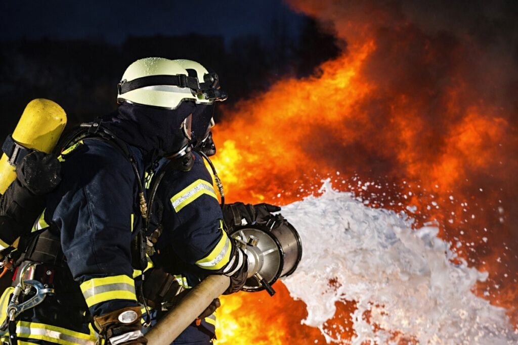 Реалистично изображение на пожарникари в пълно защитно оборудване, които гасят интензивен пожар с пожарогасителна пяна, на фона на високи пламъци и гъст дим, заснето в нощна обстановка с хоризонтална композиция.