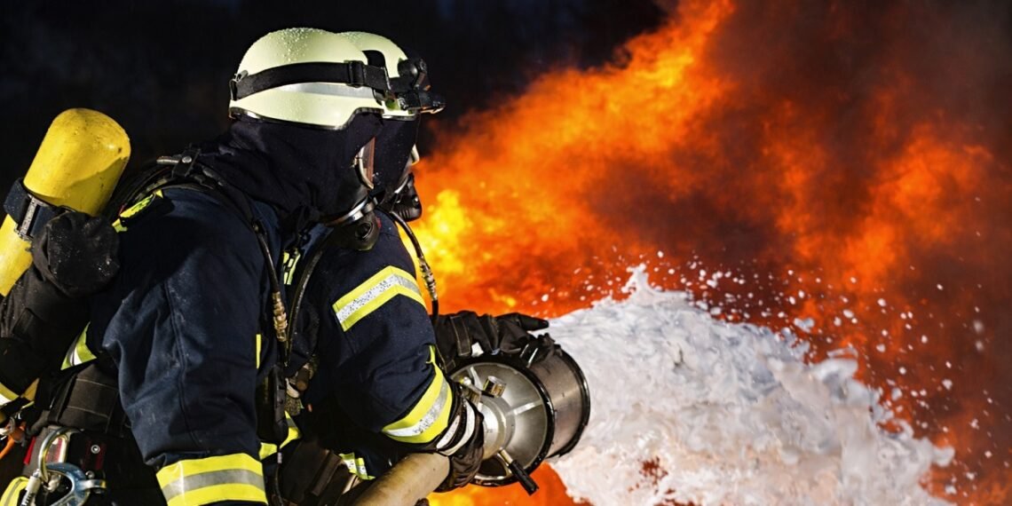 Реалистично изображение на пожарникари в пълно защитно оборудване, които гасят интензивен пожар с пожарогасителна пяна, на фона на високи пламъци и гъст дим, заснето в нощна обстановка с хоризонтална композиция.
