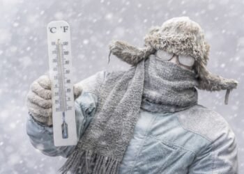 Човек в дебело зимно облекло, покрит със сняг, държи термометър с отрицателни температури по време на силен снеговалеж, заснет хоризонтално в студена зимна обстановка.