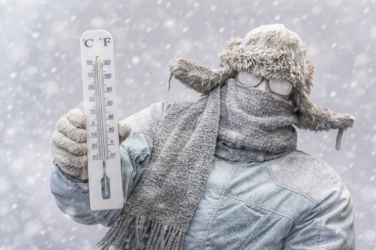 Човек в дебело зимно облекло, покрит със сняг, държи термометър с отрицателни температури по време на силен снеговалеж, заснет хоризонтално в студена зимна обстановка.