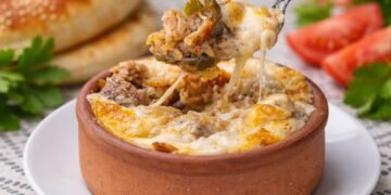 Турски харем гювеч с телешко месо, арпа шехрие и зеленчуци, покрит с разтопен бешамел и кашкавал, поднесен в глинено гювече с топла питка на заден план