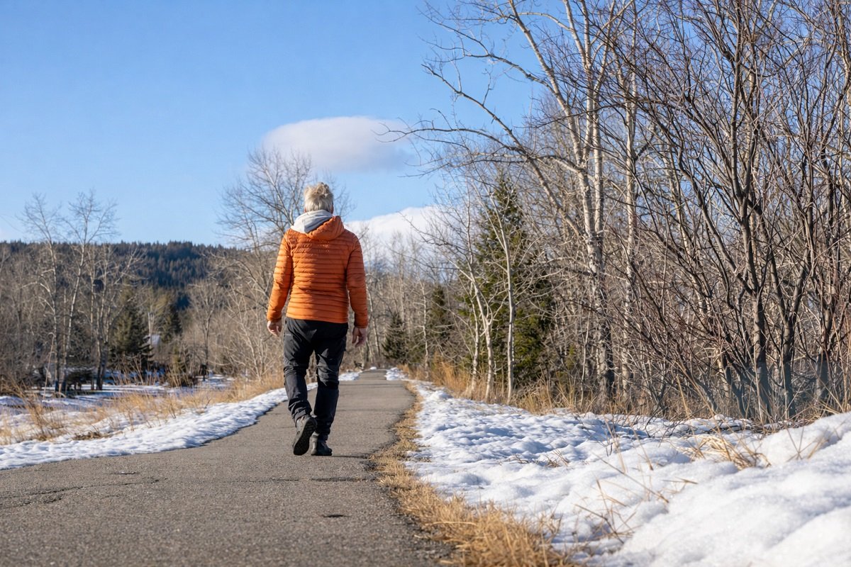 Човек с оранжево яке върви по асфалтирана алея сред зимен пейзаж с остатъци от сняг, голи дървета и ясно синьо небе, гледка отзад, хоризонтален кадър.
