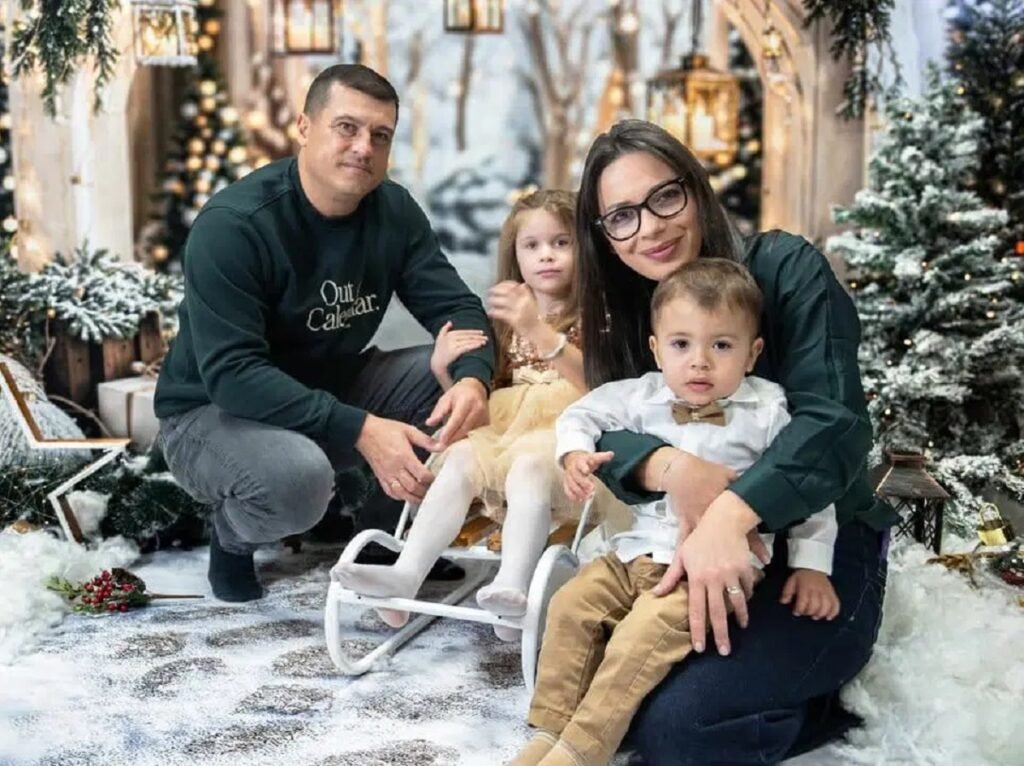 Младо семейство с две малки деца позира в зимна коледна обстановка с елхи, лампички и декоративен сняг.