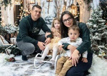 Младо семейство с две малки деца позира в зимна коледна обстановка с елхи, лампички и декоративен сняг.