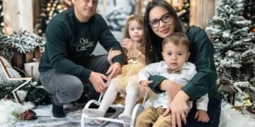 Младо семейство с две малки деца позира в зимна коледна обстановка с елхи, лампички и декоративен сняг.