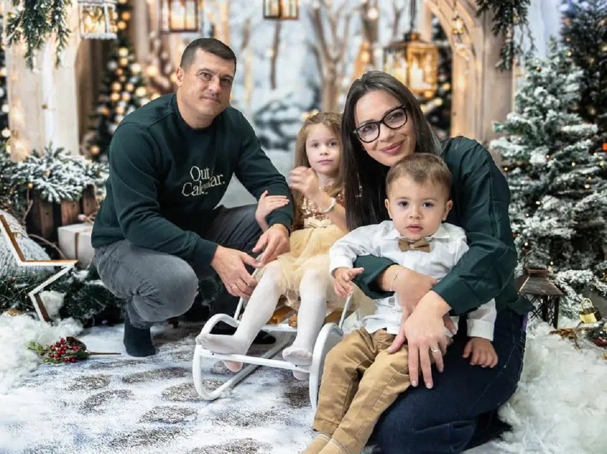 Младо семейство с две малки деца позира в зимна коледна обстановка с елхи, лампички и декоративен сняг.